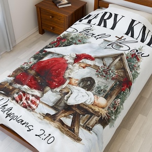 May include: White blanket with a Christmas scene depicting Santa Claus kneeling before baby Jesus in a manger. The blanket has the text "Every Knee Shall Bow" and "Philippians 2:10". The blanket is on a bed.