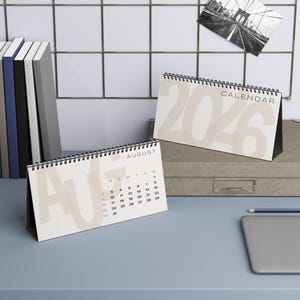 Könnte beinhalten: Zwei Tischkalender in neutralen Farbtönen. Einer zeigt den August, der andere das Jahr 2026. Beide sind spiralgebunden und stehen auf einem Schreibtisch. Ein Bleistift und ein Laptop sind ebenfalls zu sehen.