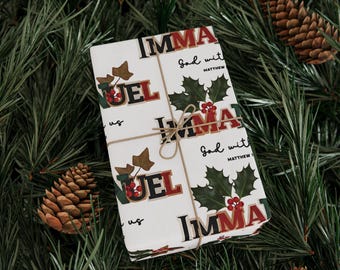 May include: A wrapped gift with a white background featuring the words "IMMA" and "NOEL" in red and black lettering, holly leaves, and berries. The gift is tied with twine and rests on a bed of pine needles with pine cones.