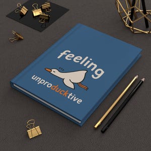 May include: A blue journal with the words "feeling unproductive" and a cartoon duck illustration. The journal is surrounded by gold binder clips, pencils, and decorative objects on a dark gray surface.