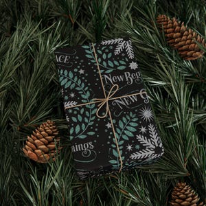 May include: A wrapped gift with a black background, adorned with teal and white botanical and star patterns, and the words "New Beginnings" and "Peace". Tied with twine, it rests on a bed of green pine needles and pine cones.