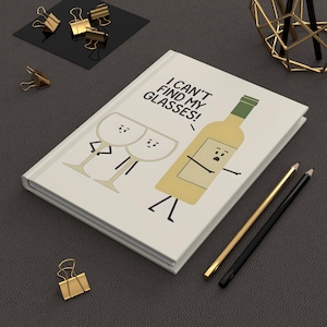 May include: A cream-coloured journal featuring the phrase "I can't find my glasses!" alongside cartoon wine glasses and a bottle. Gold binder clips and pencils are also visible.