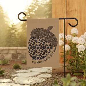May include: A tan garden flag with an acorn design. The top of the acorn is brown with white spots, and the bottom has a leopard print. The text "I'M NUTS ABOUT AUTUMN" is printed on the flag. The flag is displayed outdoors.