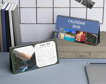 Könnte beinhalten: Zwei Schreibtischkalender für 2026. Ein Kalender hat ein blaues Cover mit dem Text "CALENDAR 2026" und vier saisonalen Bildern. Der andere Kalender hat ein grünes Cover mit dem Text "August | 2026" und einem malerischen Wasserfallbild. Ein Schwarz-Weiß-Foto einer Brücke hängt an der Wand.