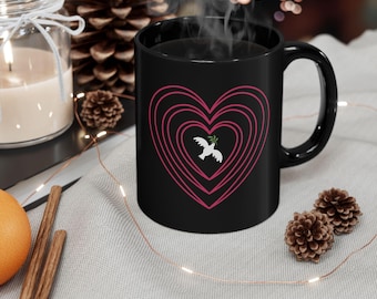 May include: A black ceramic mug featuring a pink heart design with a white dove holding an olive branch. The mug is filled with a dark liquid, possibly coffee or tea. The mug is on a light gray surface with decorative elements.