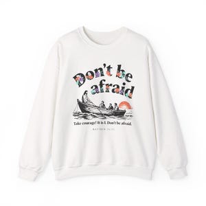 Könnte beinhalten: Ein weißes Sweatshirt mit Rundhalsausschnitt und dem Schriftzug "Don't be afraid" in schwarzer Schrift, verziert mit floralen Designs. Darunter befindet sich eine Schwarz-Weiß-Illustration von Personen in einem Boot mit dem Text "Take courage! It is I. Don't be afraid, Matthew 14:22."