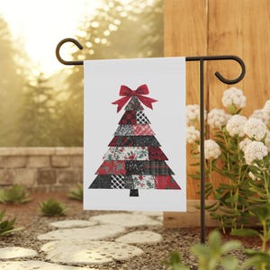 May include: A white garden flag featuring a patchwork Christmas tree design. The tree is composed of various red, black, and white patterned fabrics and topped with a red bow. The flag is displayed on a black metal garden stake.
