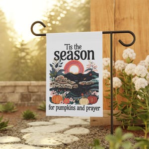 May include: A white garden flag with the text "'Tis the season" and "for pumpkins and prayer." The flag features a colorful illustration of pumpkins, mountains, and a sunset. The flag is displayed on a black metal stand.