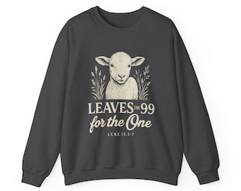Può includere: Felpa grigio scuro con un'illustrazione color crema di un agnello. Il testo recita "LEAVES the 99 for the One" e Luca 15:3-7. Il design include steli simili al grano su entrambi i lati dell'agnello.