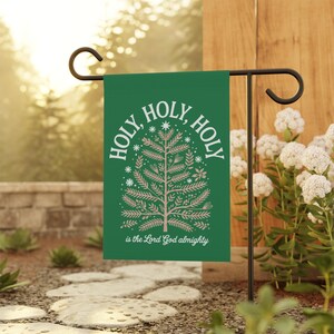 Pode incluir: Uma bandeira de jardim verde com as palavras "HOLY, HOLY, HOLY" em branco, circundando um desenho de árvore marrom claro. O texto "is the Lord God almighty" está na parte inferior. A bandeira é exibida ao ar livre em um suporte de metal preto.