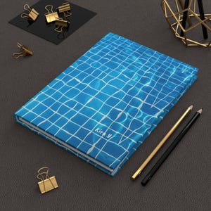 Puede incluir: Una libreta azul y blanca con un patrón de agua de piscina y el texto "Kira y" en la portada. La libreta está sobre una superficie gris oscura con dos lápices y clips de papel dorados.