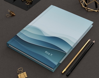 May include: A blue and white notebook with a wave pattern and the name "Kira Y." printed on the cover. The notebook is lying on a dark gray surface.