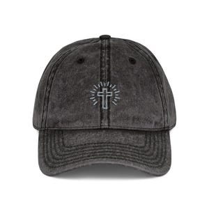 May include: A dark gray baseball cap with a curved brim. The cap features a white embroidered cross design with radiating lines. The hat has a vintage, worn look.
