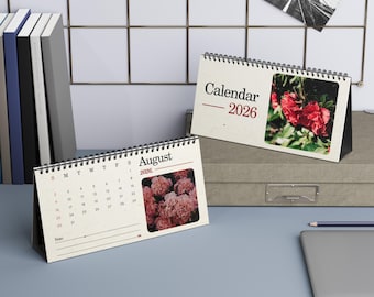 May include: Two desk calendars for 2026 are shown, one with the text "August" and a floral image, the other with "Calendar" and a red flower image. Both have a spiral binding and stand upright on a desk.