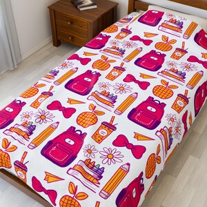 Könnte beinhalten: Weiße Bettdecke mit einem sich wiederholenden Muster von Illustrationen zum Thema Schule. Das Design umfasst rosa Rucksäcke, gelbe Bleistifte, Bücher, Gänseblümchen, Äpfel, Schleifen und Kaffeetassen. Die Bettdecke liegt auf einem Holzrahmen.