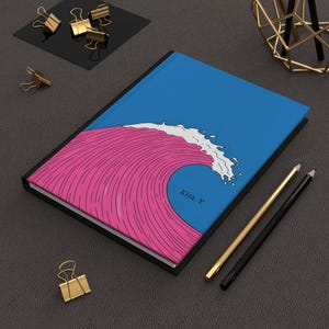 May include: A blue notebook with a pink and white wave design. The notebook has a black border and the name "Kira. Y" is printed on the front.
