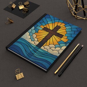 May include: A hardcover journal with a stained-glass design featuring a brown cross, yellow sun, blue sky, and white clouds. The journal is black with a lined interior. Gold and black pencils and paper clips are also visible.