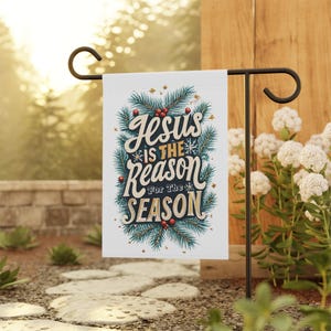Pode incluir: Uma bandeira de jardim branca com a mensagem "Jesus is the Reason for the Season" em uma fonte decorativa. O texto é cercado por galhos de pinheiro verdes, bagas vermelhas e estrelas douradas. A bandeira está em um suporte de metal preto.