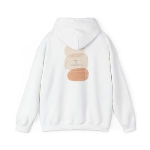 Puede incluir: Sudadera con capucha blanca con mangas largas. La parte trasera presenta un diseño abstracto de formas superpuestas en beige y naranja, con el texto "SERENITY IN SIMPLICITY". Una prenda casual y cómoda.
