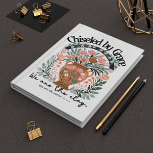 May include: White journal with the text "Chiseled by Grace" and "We are the clay, you are the potter". The cover features a floral design with a vase. Gold and black pencils and binder clips are also visible.