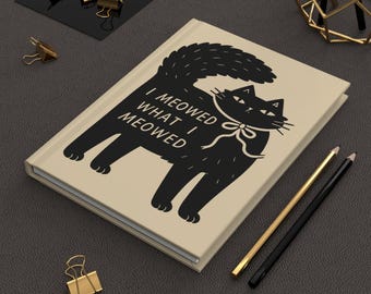 以下が含まれることがあります： 黒猫のイラストと「I meowed what I meowed」の文字が入ったベージュ色のジャーナル。猫は蝶ネクタイをしています。金と黒の鉛筆とペーパークリップも表示されています。