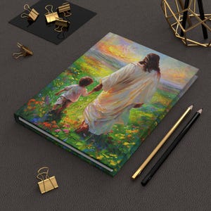 Op de afbeelding: Een hardcover notitieboek met een levendig schilderij van een figuur in witte gewaden die een kind door een kleurrijk veld leidt. Het kunstwerk heeft een zachte, impressionistische stijl met een warm kleurenpalet. Gouden en zwarte pennen en paperclips liggen in de buurt.