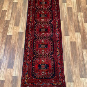 May include: A long, red runner rug featuring a repeating geometric pattern in navy, gold, and cream. The rug has a fringed edge and is displayed on a wooden floor. The rug's design is symmetrical and measures approximately 240 cm in length.