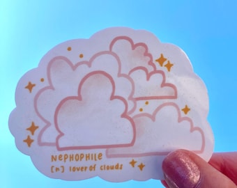 May include: A sticker with a cloud design in pink and white, with the word "NEPHOPHILE" and the definition "lover of clouds" printed on it. The sticker has a light blue background and is held by a hand.