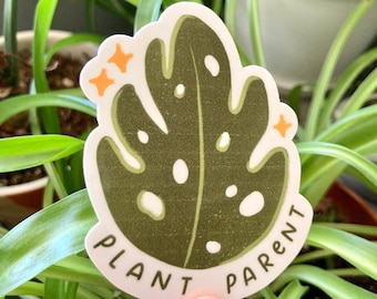 May include: A sticker featuring a green monstera leaf design with white spots and the words "PLANT PARENT" in black. The sticker has a white border and orange star accents. The sticker is held in front of a background of green plants.