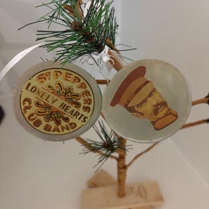 May include: Two round Christmas ornaments with a Beatles theme hang from a small artificial tree. One ornament features the text "Sgt. Pepper's Lonely Hearts Club Band". The other shows a cartoon image of a person in a hat. The tree has a wooden base.