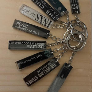 May include: A collection of rectangular keychains featuring band names and album titles, including "The Rolling Stones," "AC/DC," and "U2." The keychains are attached to a silver chain and key rings, set against a light wood background.
