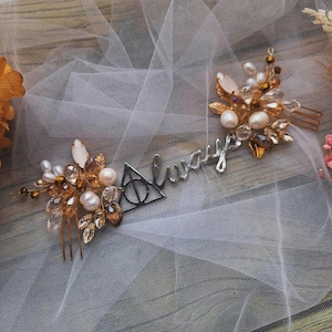 Op de afbeelding: Een zilverkleurige haarkam met het woord Always in cursief schrift, een driehoekig symbool en clusters van witte parels en heldere, amberkleurige kristallen. Het decoratieve sieraad is afgewerkt met metalen blaadjes en draadaccenten.
