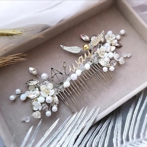 May include: A decorative hair comb with a silver metal design, featuring the word "Always" in cursive. It's embellished with white flowers, pearls, crystals, and silver wings. The design incorporates the Deathly Hallows symbol.
