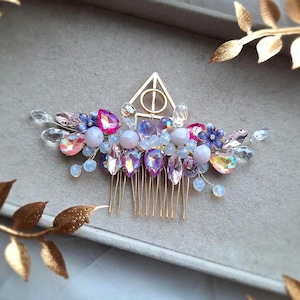 May include: A decorative hair comb with a gold-coloured triangle and circle design, embellished with pink, purple, and clear gemstones, small blue flowers, and clear beads. A beautiful hair accessory for special occasions.