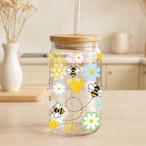May include: A clear glass drinking jar with a bamboo lid and a glass straw. The jar is decorated with yellow honeycomb shapes, bees, and flowers in white, pink, and blue. The jar is designed for cold beverages.