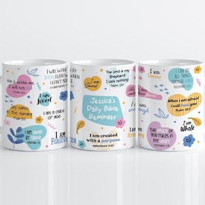 Op de afbeelding: Drie witte keramische mokken met kleurrijke tekst en illustraties. De mokken bevatten inspirerende citaten en zinnen, waaronder "I am Saved" en "I am Divine". De middelste mok leest "Jessica's Daily Bible Reminder."