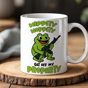 Pode incluir: Caneca de cerâmica branca com um sapo de desenho animado segurando um rifle. A caneca tem o texto "HIPPITY HOPPITY" acima do sapo e "GET OFF MY PROPERTY" abaixo. O sapo é verde com boca vermelha e rifle preto.
