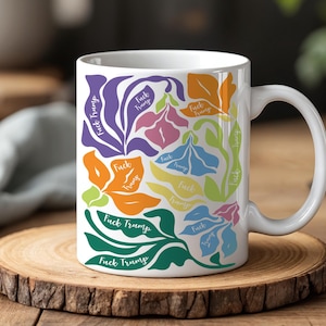 May include: White ceramic mug featuring a colorful floral design with the text "Fuck Trump" in various colors. The flowers are in shades of purple, orange, pink, blue, and green. The mug sits on a wooden coaster.