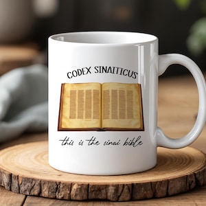 Op de afbeelding: Witte keramische mok met de tekst "CODEX SINAITICUS" en een illustratie van een open boek met verouderde pagina's. Daaronder staat de tekst "this is the sinai bible". De mok staat op een houten onderzetter.