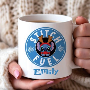 May include: White ceramic mug featuring a blue circular design with the words "Stitch Fuel" and a cartoon character wearing a reindeer antler headband. The name "Emily" is printed in blue below the design. The mug is held by a person.