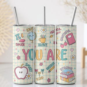 May include: Three stainless steel insulated tumblers with lids and straws. Each tumbler features a grid pattern background with the words "YOU ARE" in colorful letters. The tumblers are decorated with various illustrations and words like "brave" and "smart".