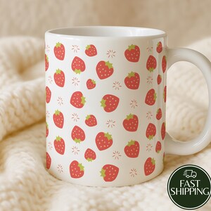Può includere: Tazza in ceramica bianca decorata con un motivo di fragole rosse con cime verdi e piccole stelle. La tazza ha un manico bianco ed è posta su uno sfondo bianco sporco.