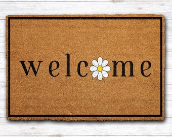 May include: A rectangular doormat with the word "welcome" in black letters. The "o" in welcome is replaced with a white daisy with a yellow center. The mat has a brown background and a black border.
