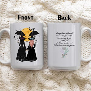 May include: A white ceramic mug showing a front and back view. The front features two figures in black dresses and hats holding umbrellas under a yellow moon. The back displays text: Always throw spilled salt over your left shoulder, keep rosemary by your garden gate, plant lavender, for luck, fall in love whenever you can, with a small illustration of herbs.