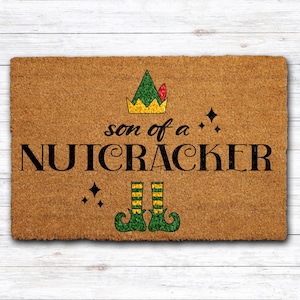 May include: A rectangular doormat with the phrase "son of a NUTCRACKER" in black lettering. The design includes a green and gold crown, elf shoes, and star accents. The mat is brown with a textured surface.
