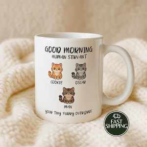 May include: A white ceramic mug featuring the black text Good Morning Human Servant at the top and Your Tiny Furry Overlords at the bottom. Three cartoon cats are illustrated in the center, labeled Cookie, Oscar, and Max in black text.