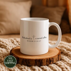May include: A white ceramic mug featuring the black printed text Motherhood is a sacred calling with a small cross icon and the reference WISDOM 31:25-27. The mug has a smooth finish and a curved handle, standing on a circular wooden slice.