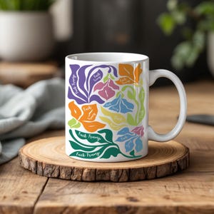 May include: A white ceramic mug featuring a colorful abstract floral pattern in shades of purple, orange, pink, blue, yellow, and green. The design includes the repeated text "Fuck Trump" integrated into the organic shapes of the flowers and leaves.
