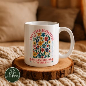 May include: A white ceramic mug featuring a colorful floral illustration inside an arch. The text reads MOTHERHOOD IS KINGDOM WORK Prov 31:25-27, with additional small text on the sides. The design includes red, blue, and yellow flowers.