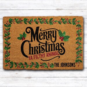 May include: A rectangular doormat with the words "Merry Christmas Ya Filthy Animal" in black script. The doormat is framed with a green and red holly berry design. The family name "The Johnsons" is printed at the bottom.